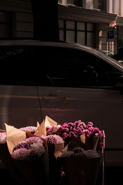 A collection of fresh flower bouquets featuring pink chrysanthemums and carnations, wrapped in brown kraft paper, are arranged in a basket outside on a city sidewalk during daylight. The flowers appea