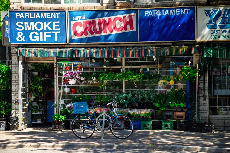 A flower shop storefront with a blue awning displaying signs that read 'Parliament Smoke & Gift' and 'Nestlé Crunch.' The open space showcases a variety of fresh flowers including pink, purple, and g