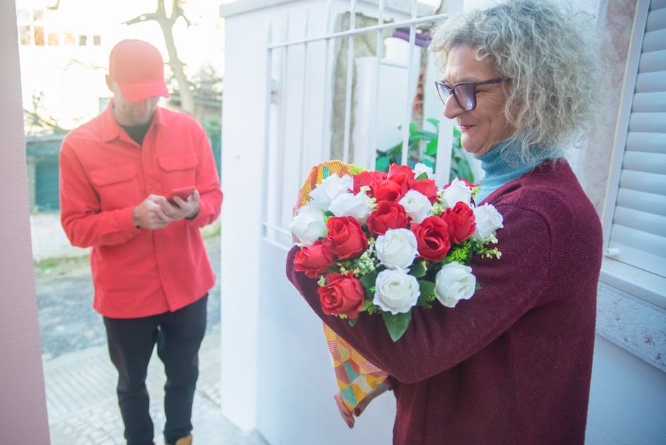 A mature woman with curly gray hair and glasses holds a vibrant bouquet of fresh red and white roses, wrapped in colorful paper, as she receives a flower delivery from a courier at her doorstep. The w