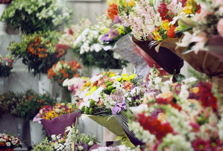 A vibrant display of floral arrangements featuring a variety of fresh flowers such as carnations, lilies, daisies, and roses in shades of pink, white, yellow, orange, and purple. The bouquets are wrap